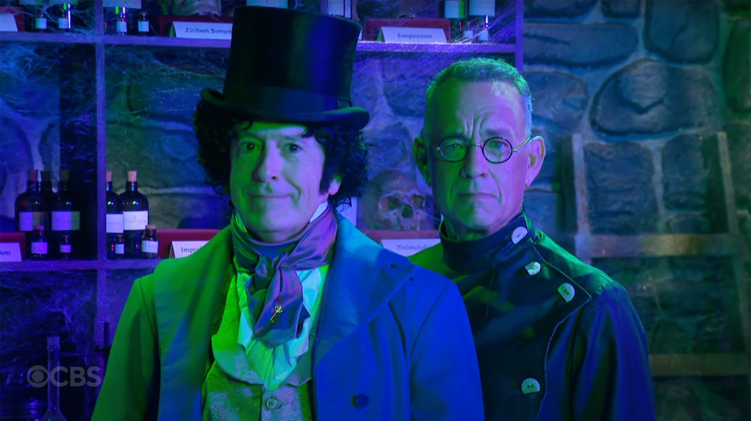 Two men in costume stand in a green lit room, staring at the camera.