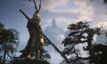 A warrior in ornate armor stands on a rocky cliff, holding a large staff, facing a colossal stone statue of a deity in the distance, surrounded by mist and trees. The scene is set against a cloudy sky with birds flying overhead.