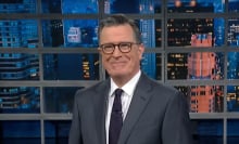 A man in a suit stands on a talk show stage, smiling mischeviously.