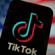 An phone displaying the TikTok logo in front of a blurry American flag.