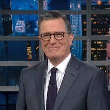 A man in a suit stands on a talk show stage, smiling mischeviously.