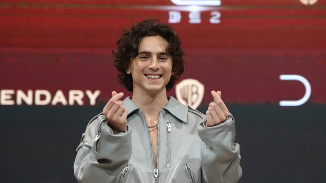 Timothée Chalamet smiling in a gray jumpsuit doing finger hearts.