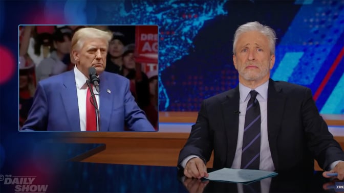 A man sits behind a talk show desk looking disappointed. In the top left is an image of another man standing at a podium, surrounded by a crowd.